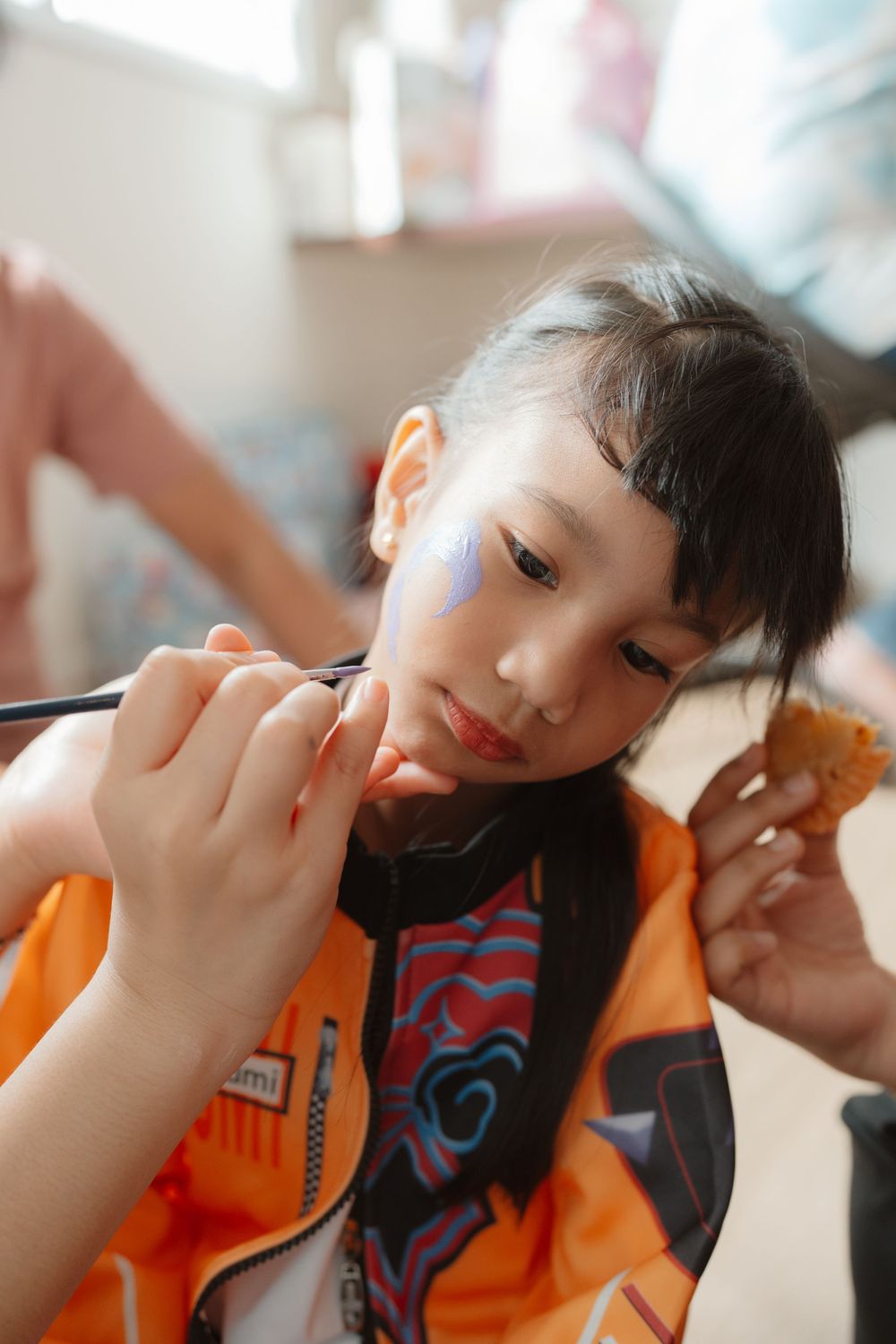 A young Asian girl in an orange shirt gets her face painted with blue design at a party event.
