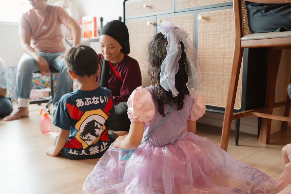 Woman sitting on floor with two children, one in a pink tutu dress, in a cozy indoor setting.