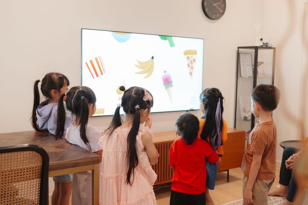 Children watching an educational food-themed video on a large TV screen in a bright classroom setting.