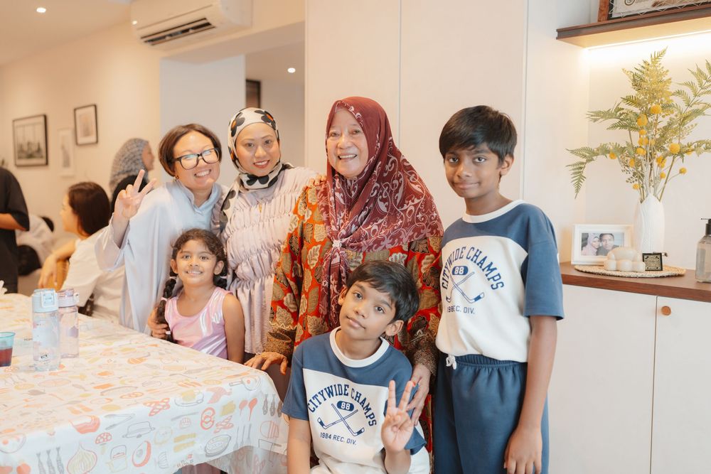 Multi-generational Muslim family posing together indoors, wearing traditional and casual clothing, smiling warmly at a gathering.