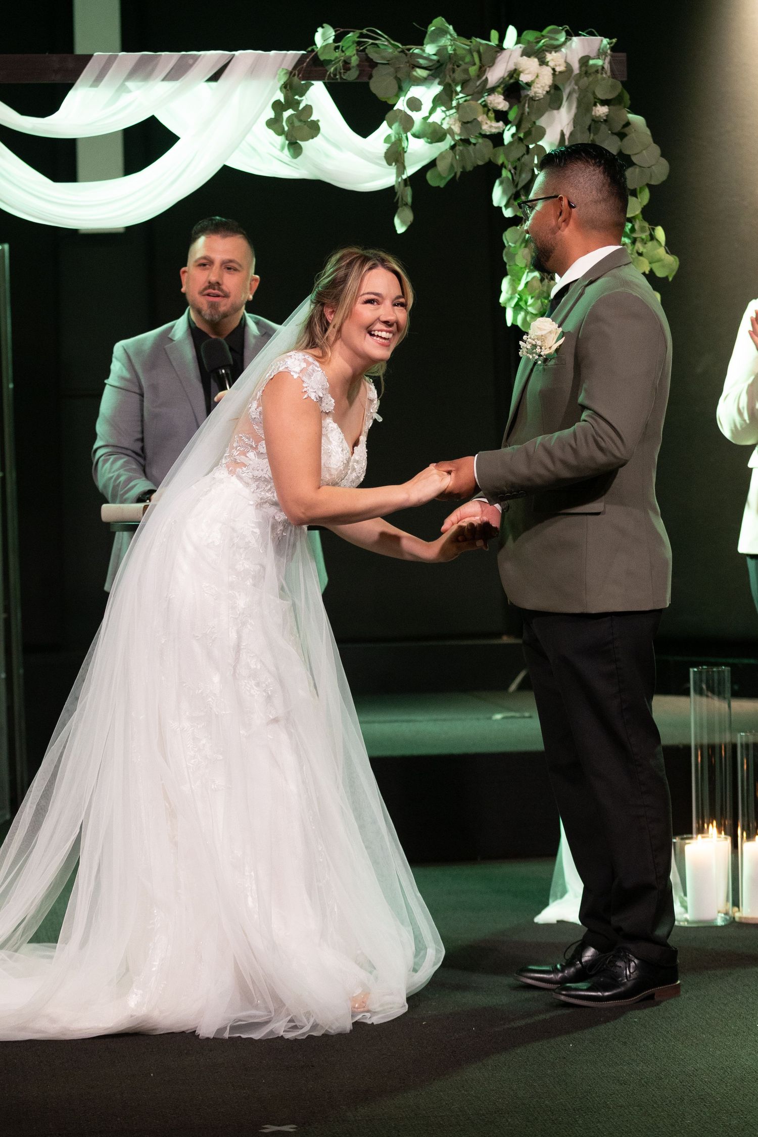 Wedding ceremony on stage with bride, groom, officiant, and groomsmen standing before floral arch with candles.