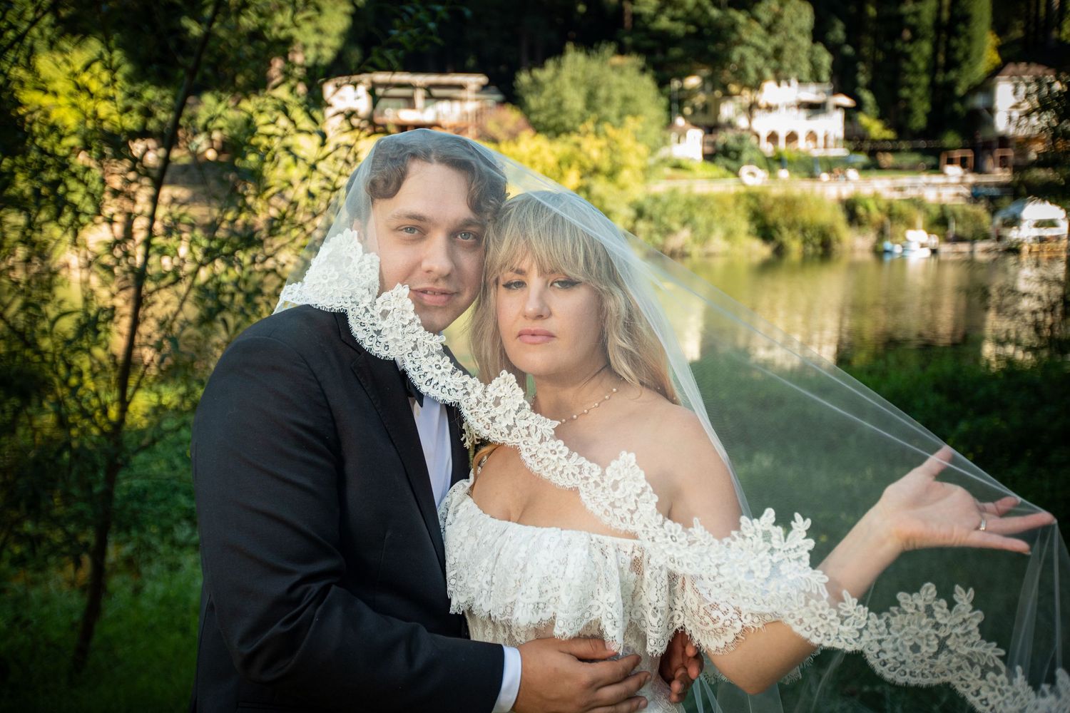 Bride and groom pose together outdoors near a lake, bride wearing lace veil and off-shoulder gown, groom in black suit.