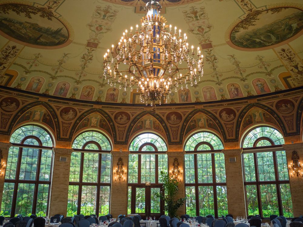 Opulent ballroom with grand chandelier, painted dome ceiling, arched windows, and elegant dining tables.