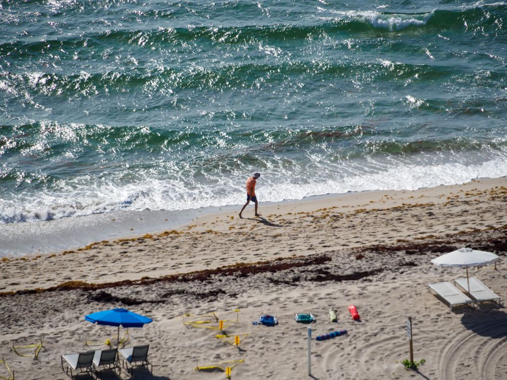 Person walking alone on a sandy beach with crashing waves and beach chairs in background.