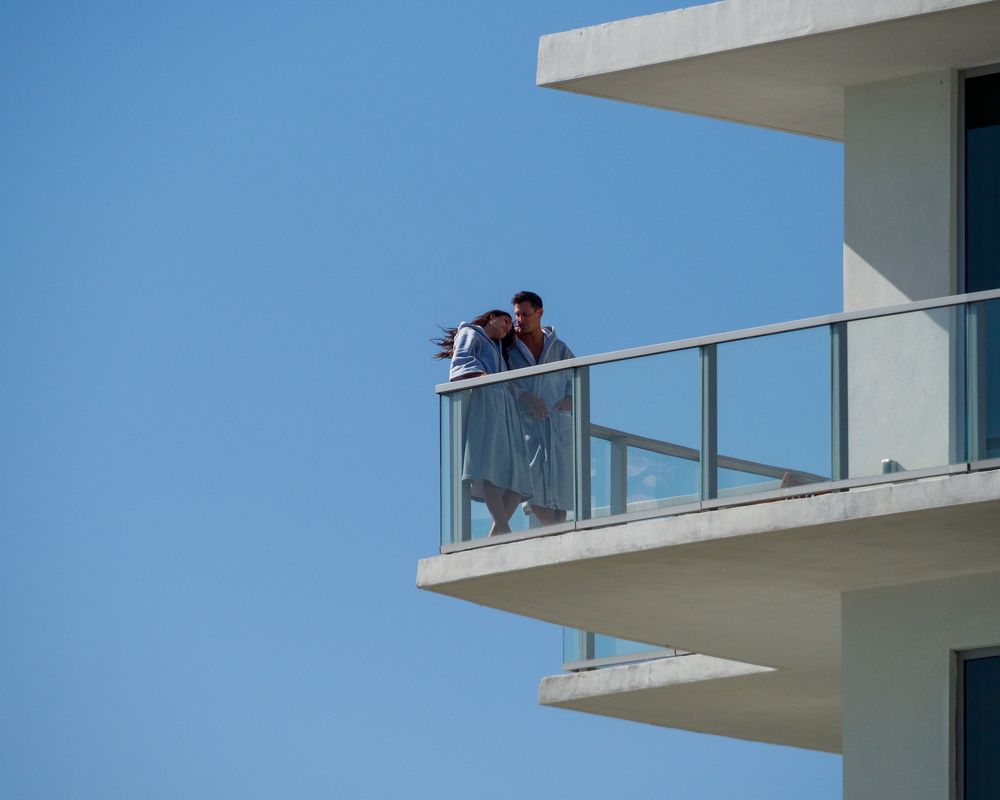Two people standing together on a modern building balcony against a clear blue sky.