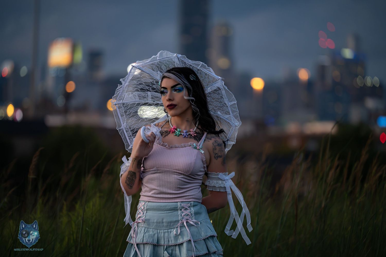 Woman in white parasol and ruffled skirt poses at dusk with city skyline backdrop in dramatic editorial fashion shoot.