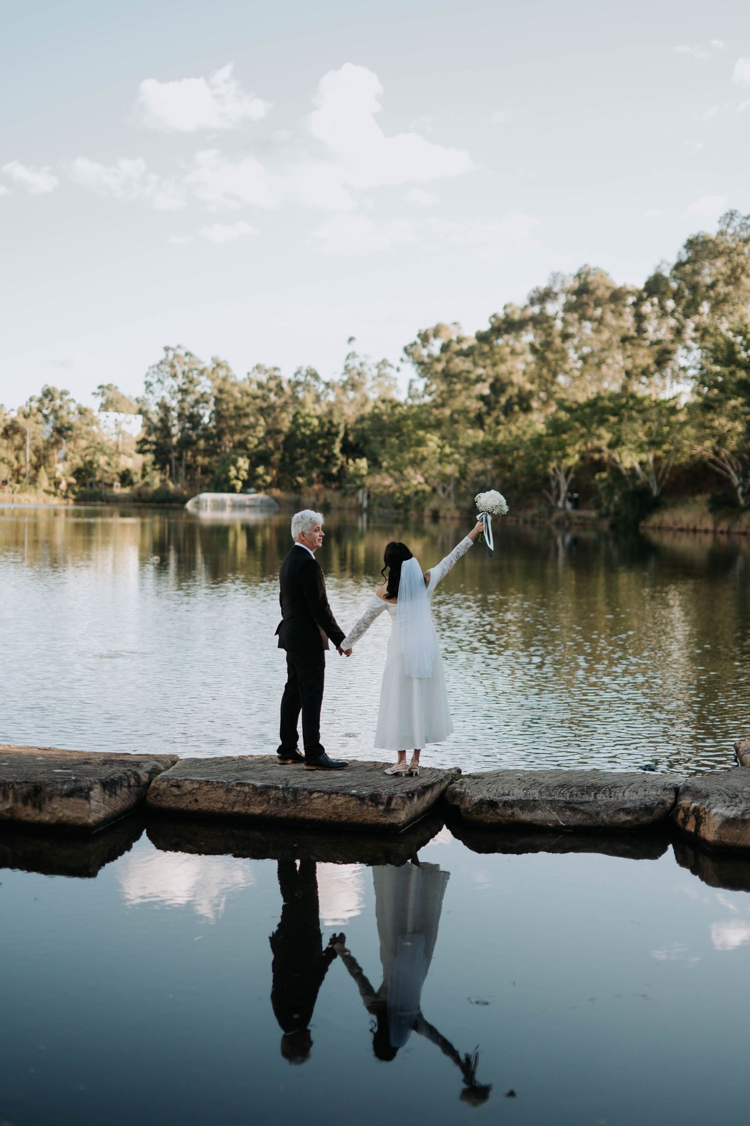 Bride and groom holding hands on stone pier by reflective lake, surrounded by lush trees, bride raising bouquet.