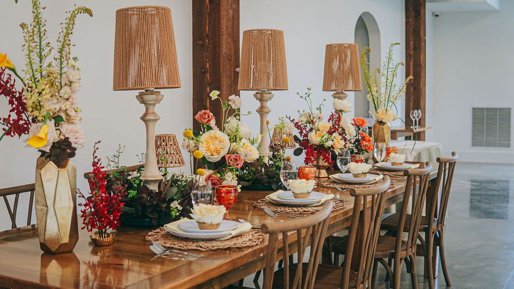 Elegant wooden dining table with colorful floral arrangements and tall wicker lampshades in a bright reception venue.