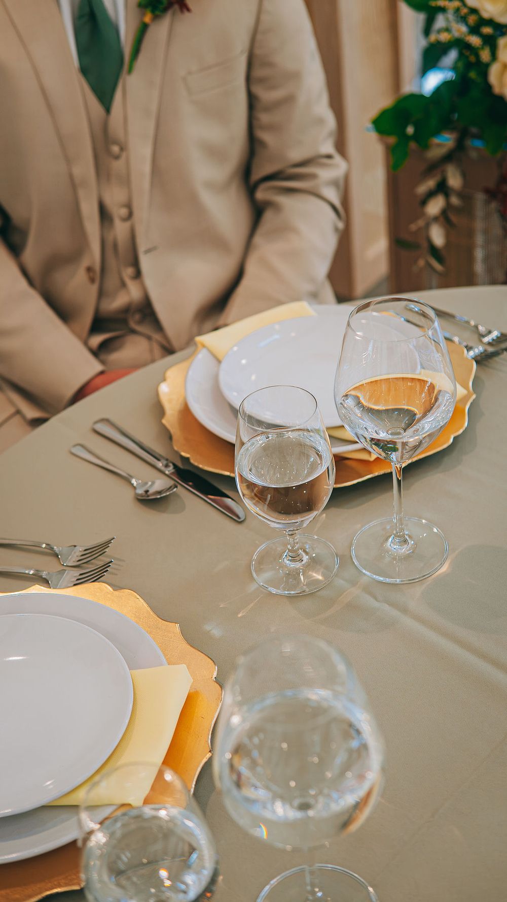 Elegant wedding table setting with gold charger plates, crystal glasses, and silverware beside a man in a beige suit.