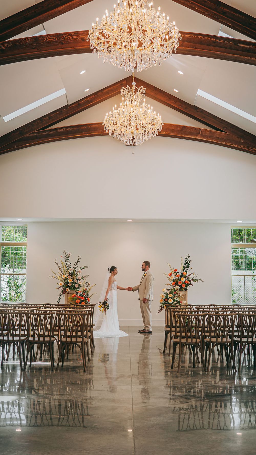 Bride and groom hold hands at altar in elegant venue with crystal chandeliers, wood beam ceiling, and floral arrangements.