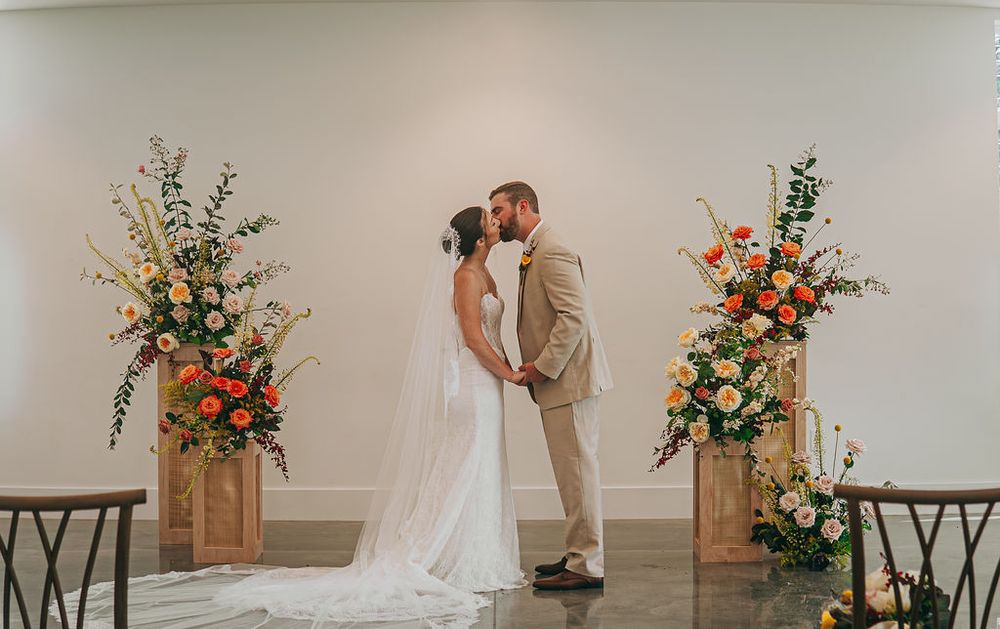 Bride and groom share first kiss at altar flanked by vibrant floral arrangements in warm tones at elegant indoor venue.
