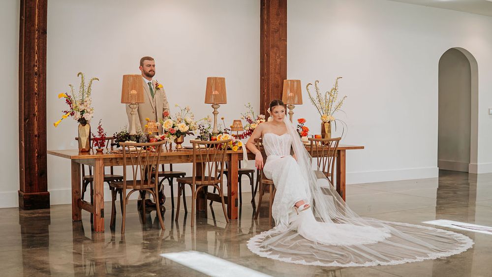 Bride and groom seated at elegant rustic wedding reception table with floral arrangements in a bright white venue.