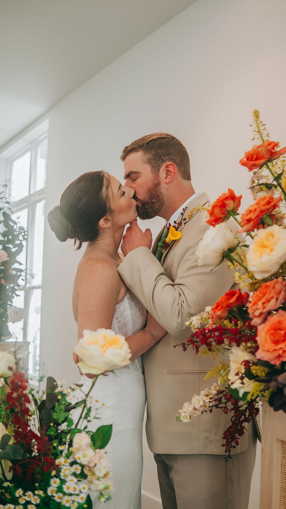 Bride and groom sharing a kiss surrounded by colorful floral arrangements during their wedding ceremony indoors.