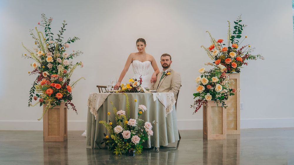Bride and groom seated at elegant sweetheart table flanked by tall wooden pedestals with colorful floral arrangements.