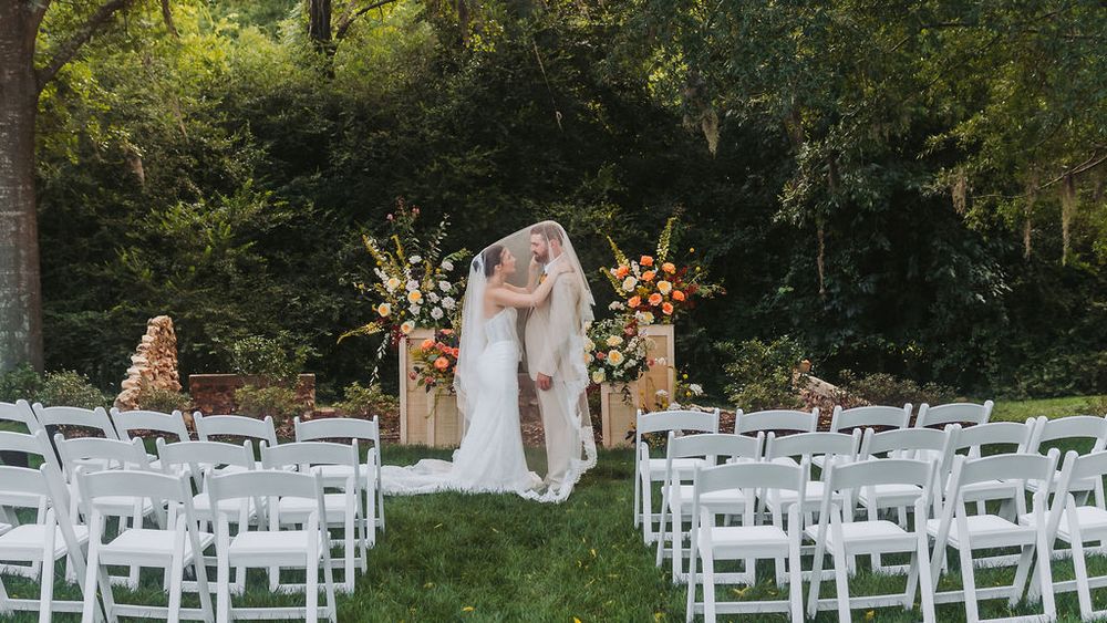 Bride and groom kissing at outdoor wedding altar with white chairs and lush greenery backdrop.