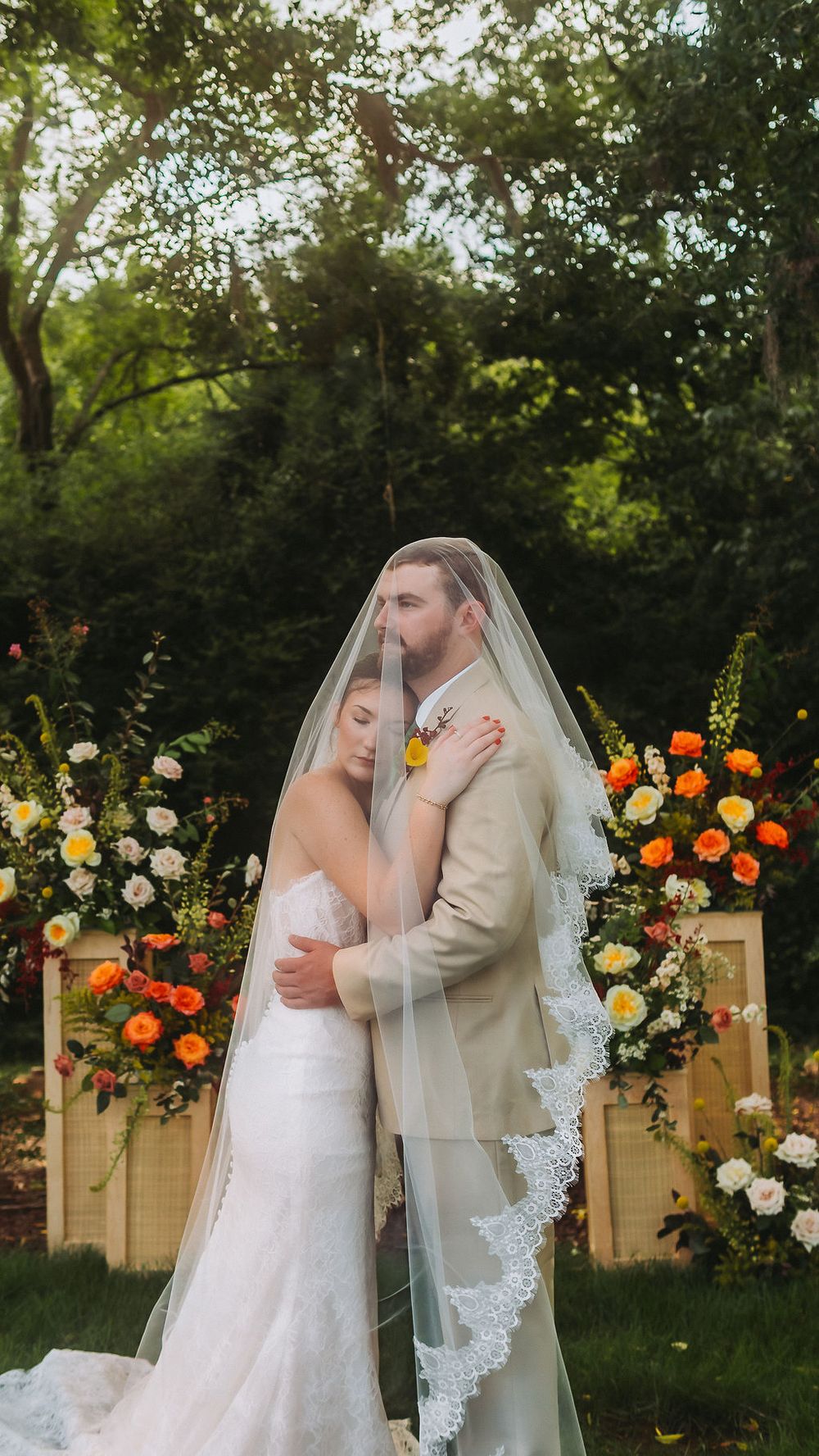 Bride and groom embrace under lace veil at outdoor wedding ceremony with colorful floral arch backdrop.