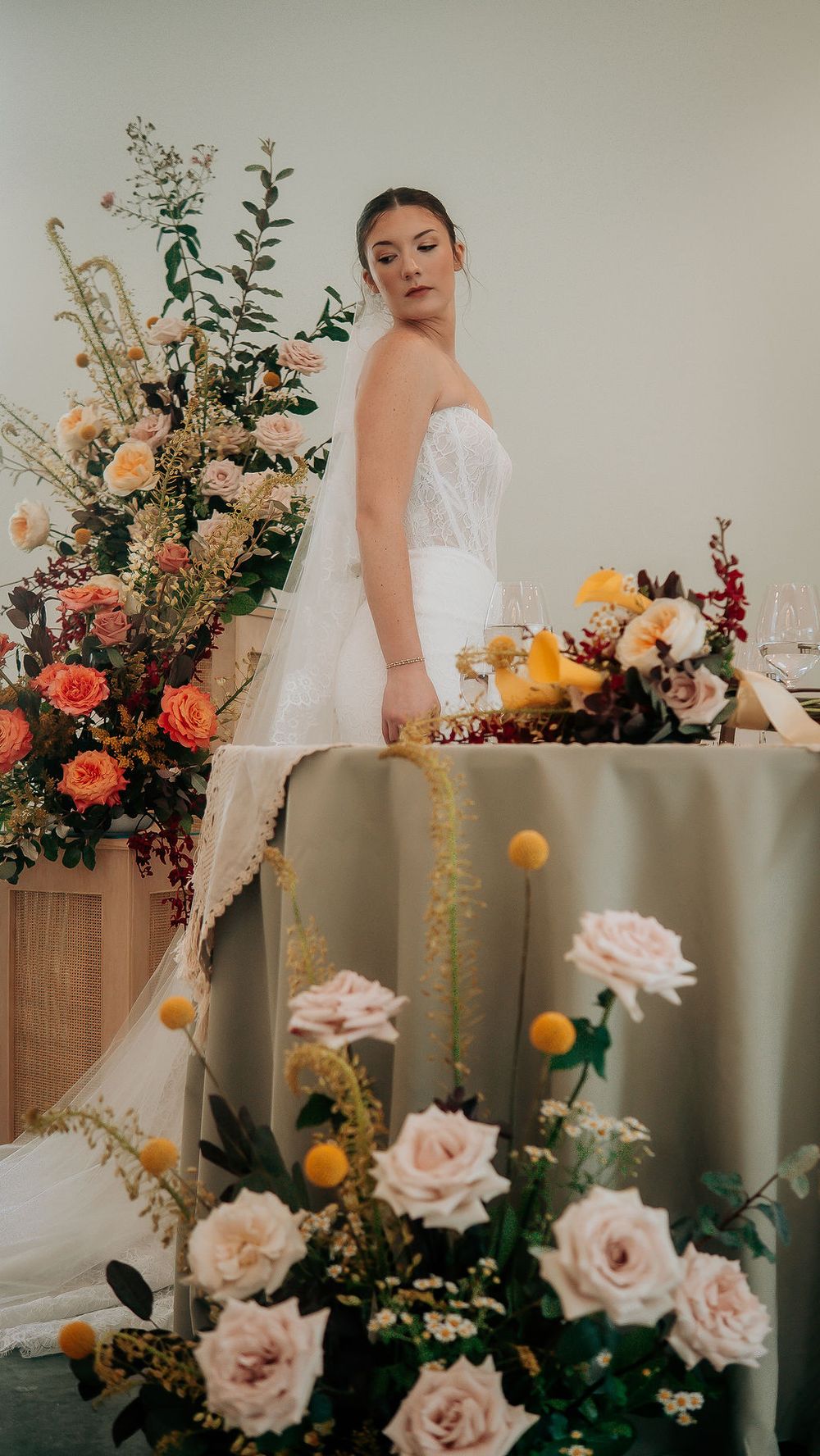 Bride in strapless white gown standing beside lush floral wedding table arrangements with roses.