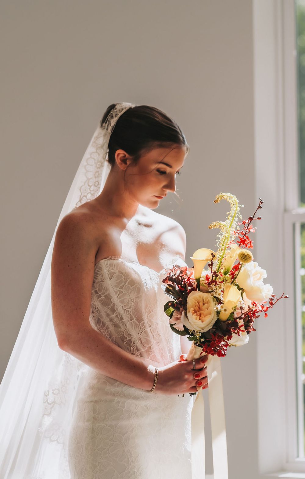 Elegant bride in lace wedding dress holding a colorful bouquet near a sunlit window.