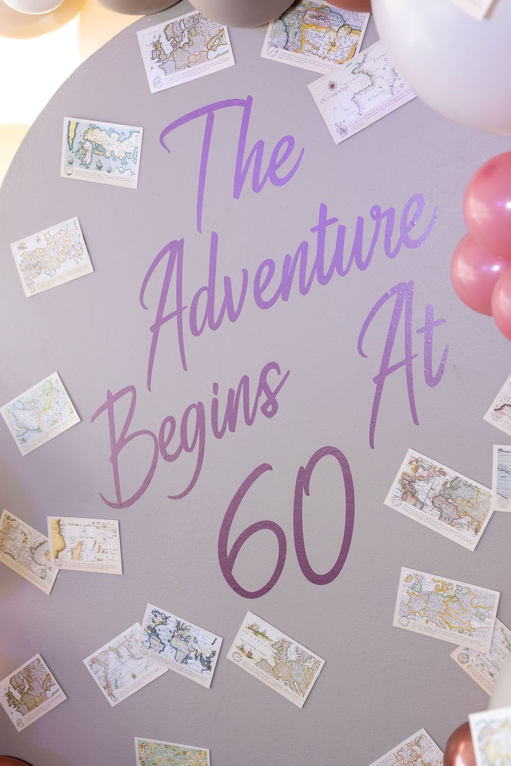 A lavender oval sign reading The Adventure Begins At 60 decorated with vintage map postcards and pink balloons for a birthday party.