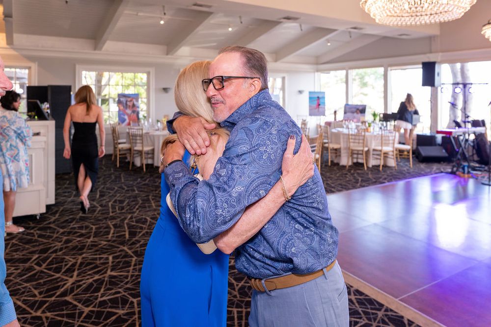 Two people share a warm hug on a dance floor at an elegant indoor event venue with chandeliers.