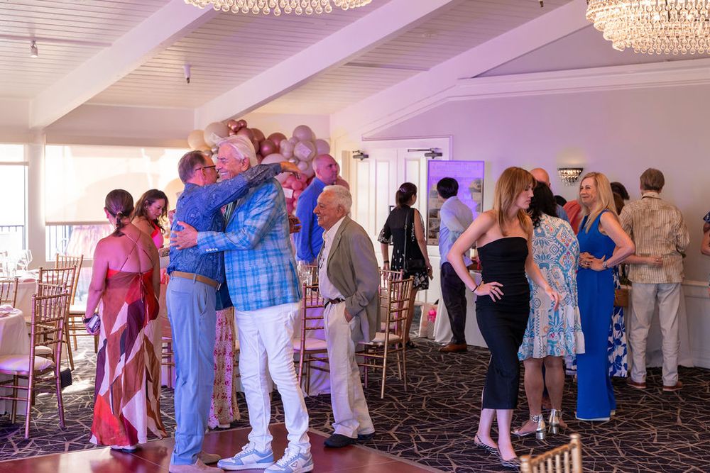 Guests mingle and dance at an elegant indoor event venue with chandeliers and large windows.