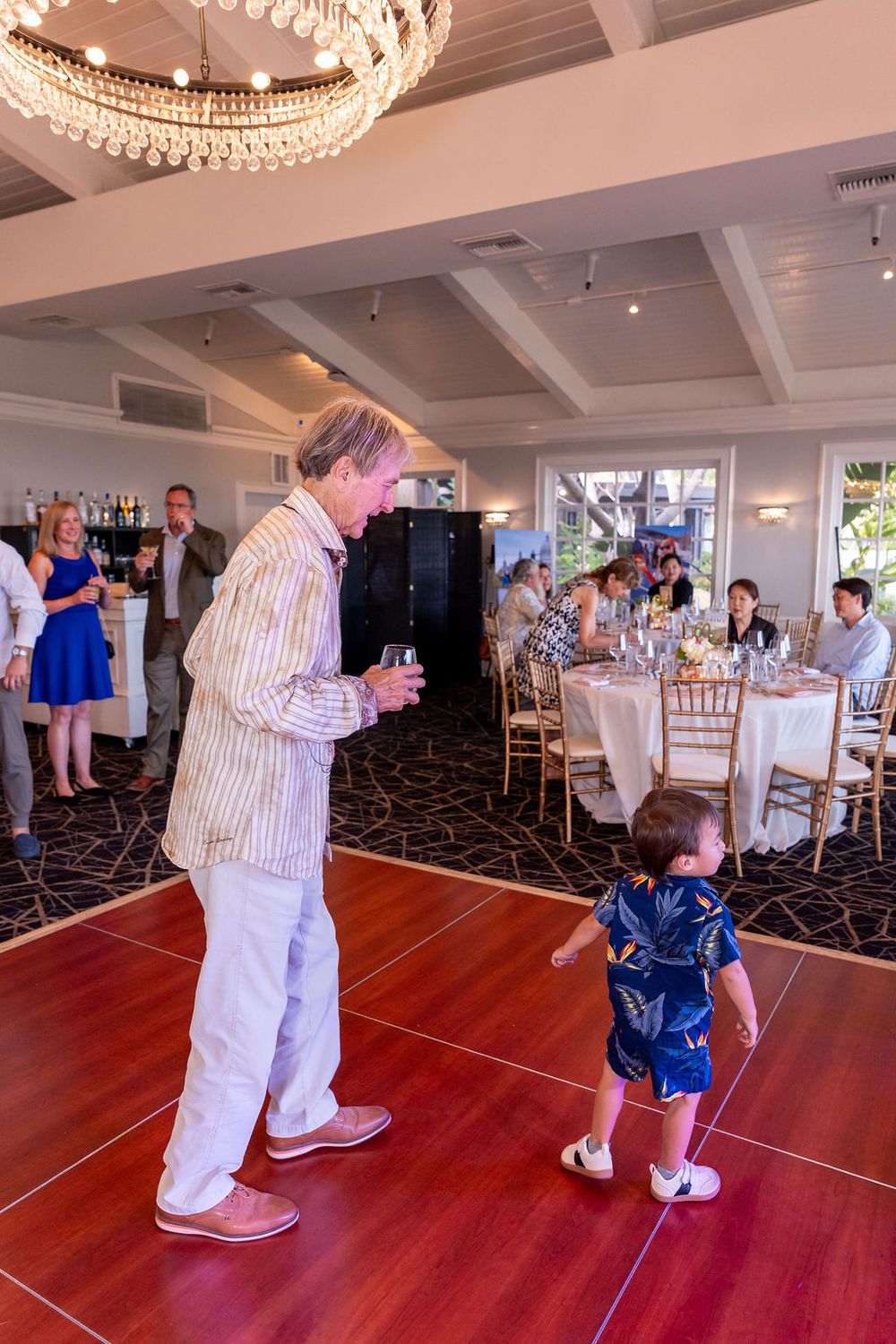 Man in striped blazer interacts with toddler on red dance floor at elegant indoor reception venue.