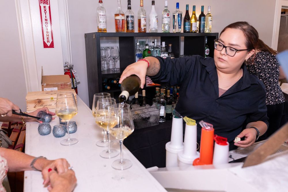 Bartender pouring white wine into glasses at a bar with bottles and supplies on display.