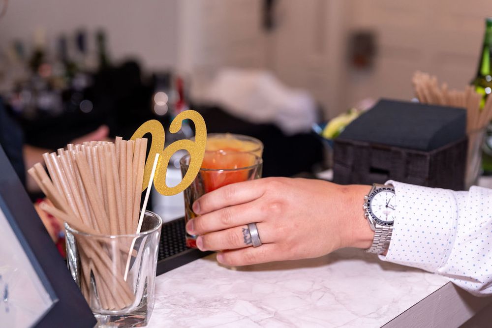 Bartender's hand holding a cocktail garnished with a gold decorative pick near straws and bar tools.