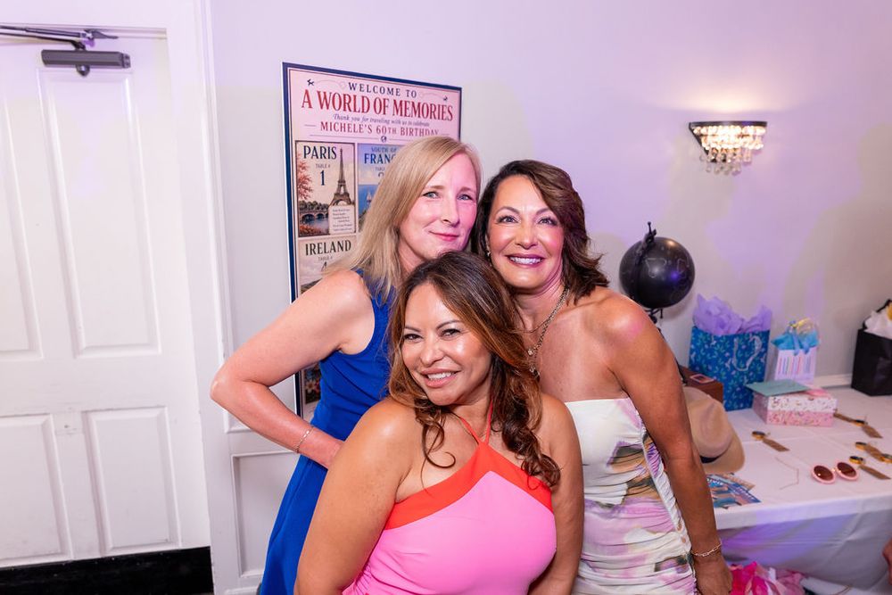 Three women smiling and posing together at an indoor celebration party with decorations in the background.