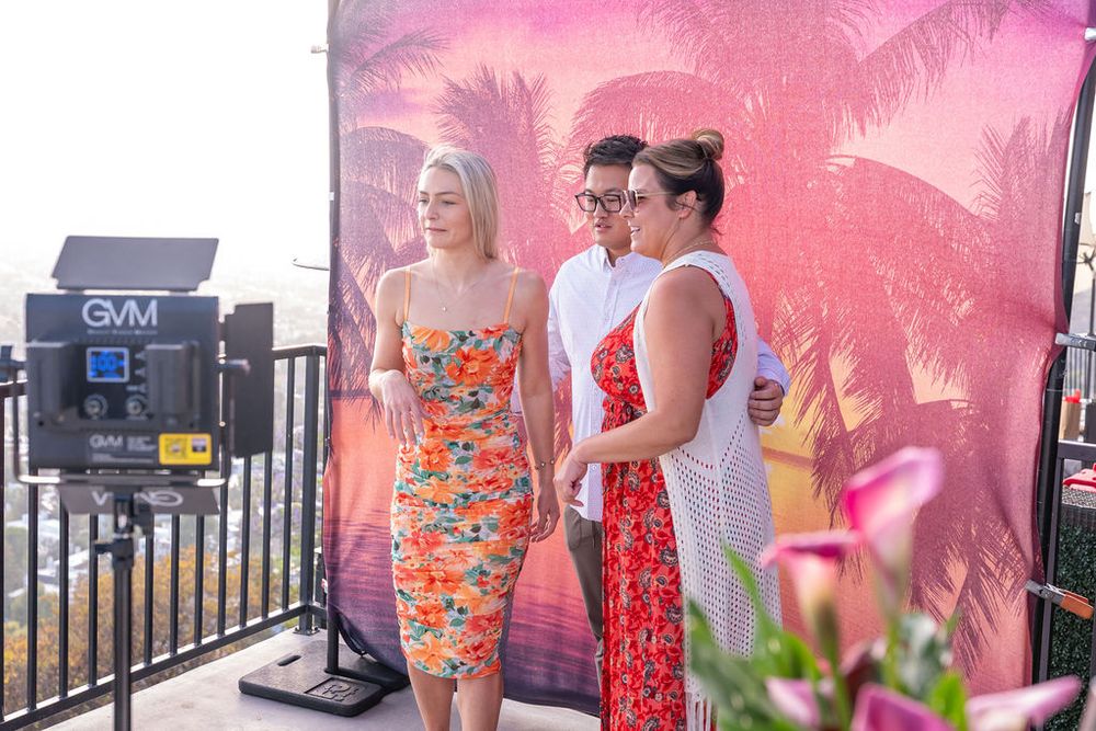 Three women in colorful dresses pose together at an outdoor event with a tropical pink palm tree backdrop.