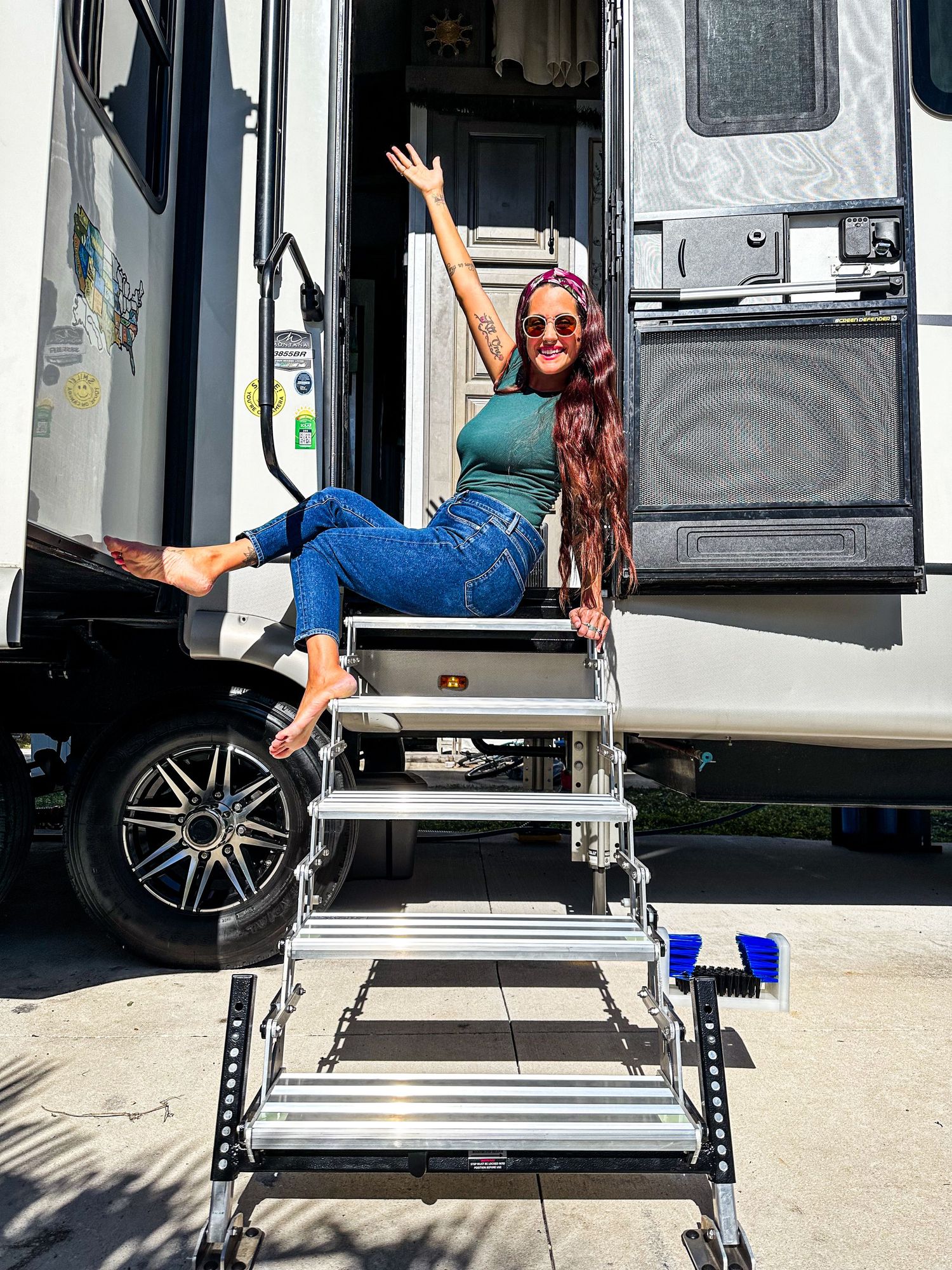 Woman with long red hair sitting on RV steps waving cheerfully outdoors.