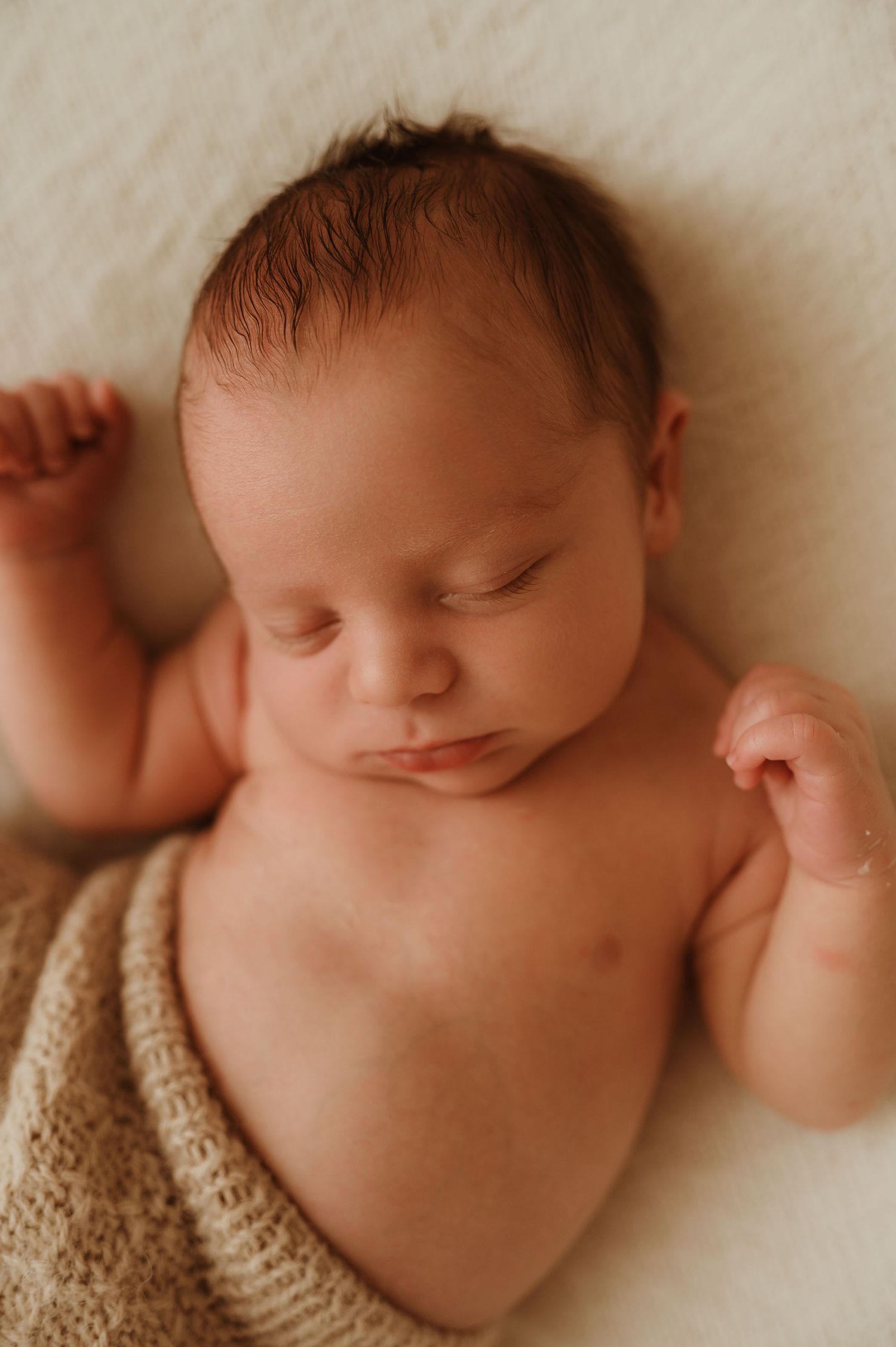 Peaceful sleeping newborn baby wrapped in a soft tan knit blanket with tiny fists raised on a white background.