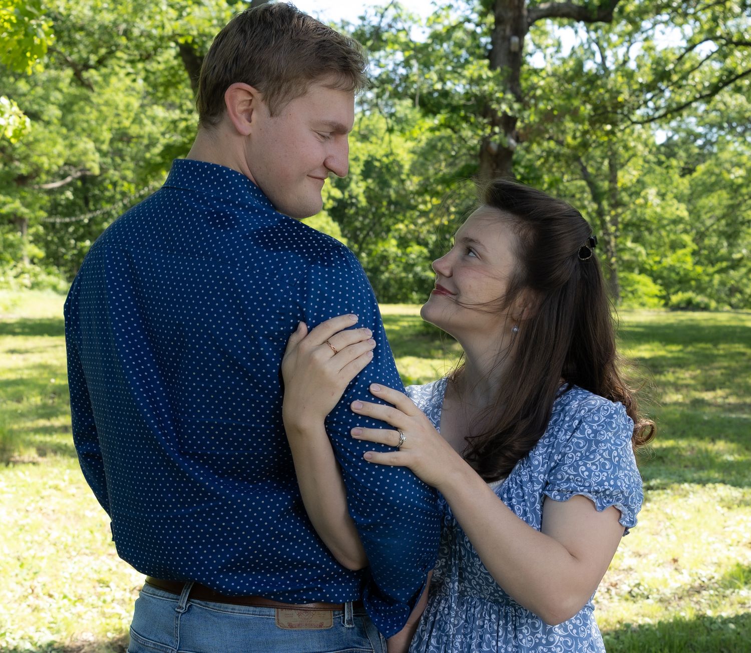 A couple shares a romantic moment outdoors, the man in a blue shirt and woman in a floral dress embracing in a green park.