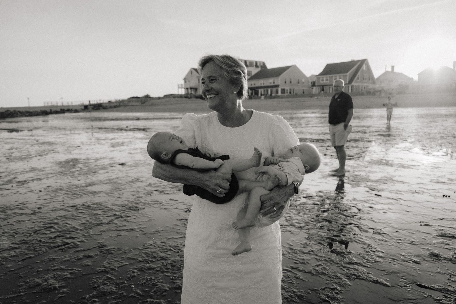 Black and white beach photo of smiling woman holding two newborn babies with coastal houses in background at sunset.
