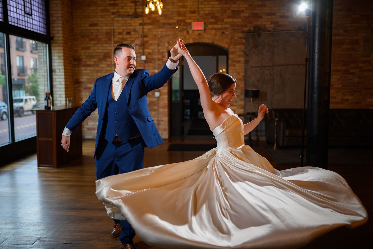 Bride and groom share a joyful first dance, her white gown swirling, in a warmly lit brick-walled wedding venue.