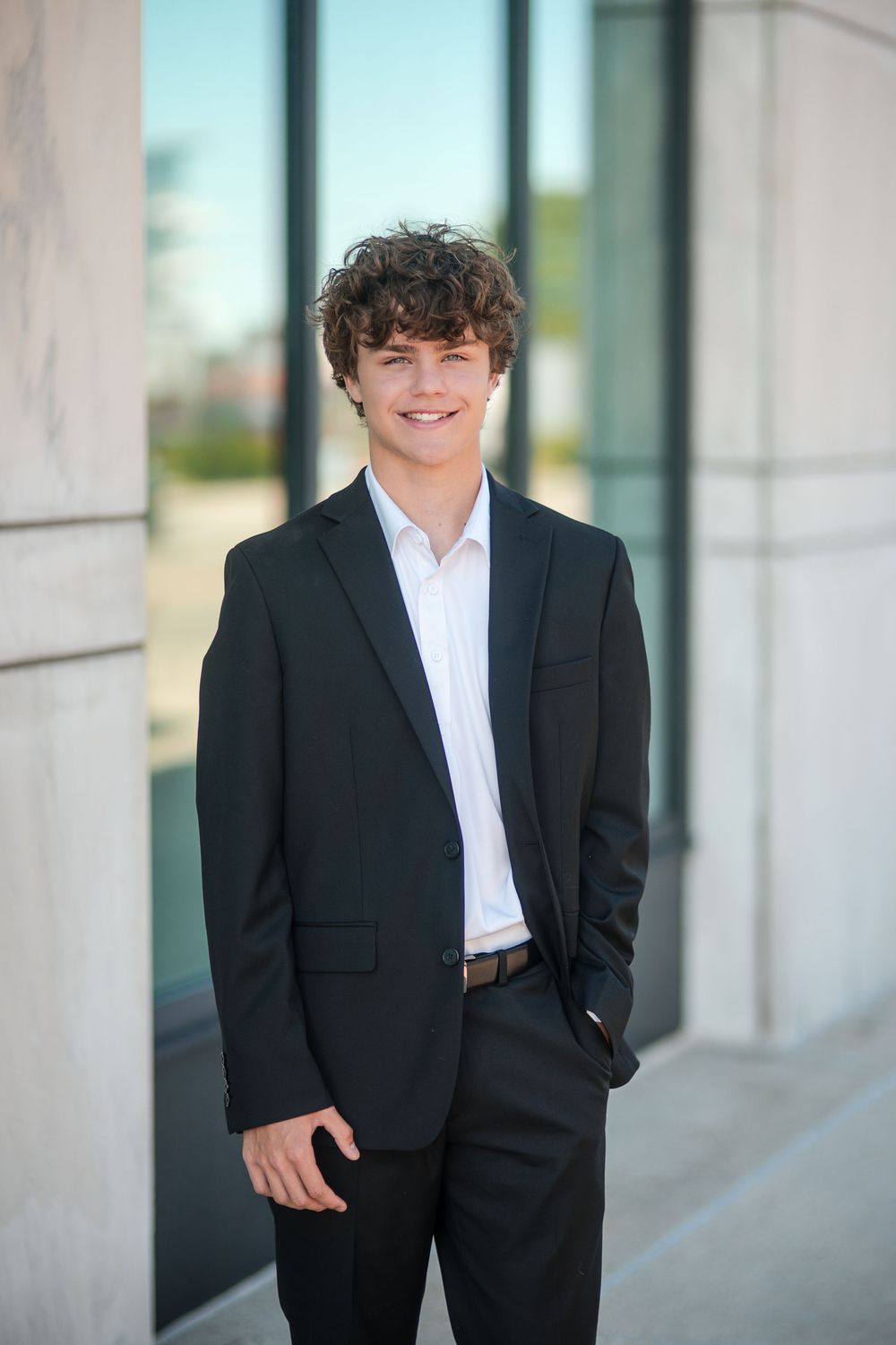 Young man in black suit and white shirt smiling outdoors near modern building columns.