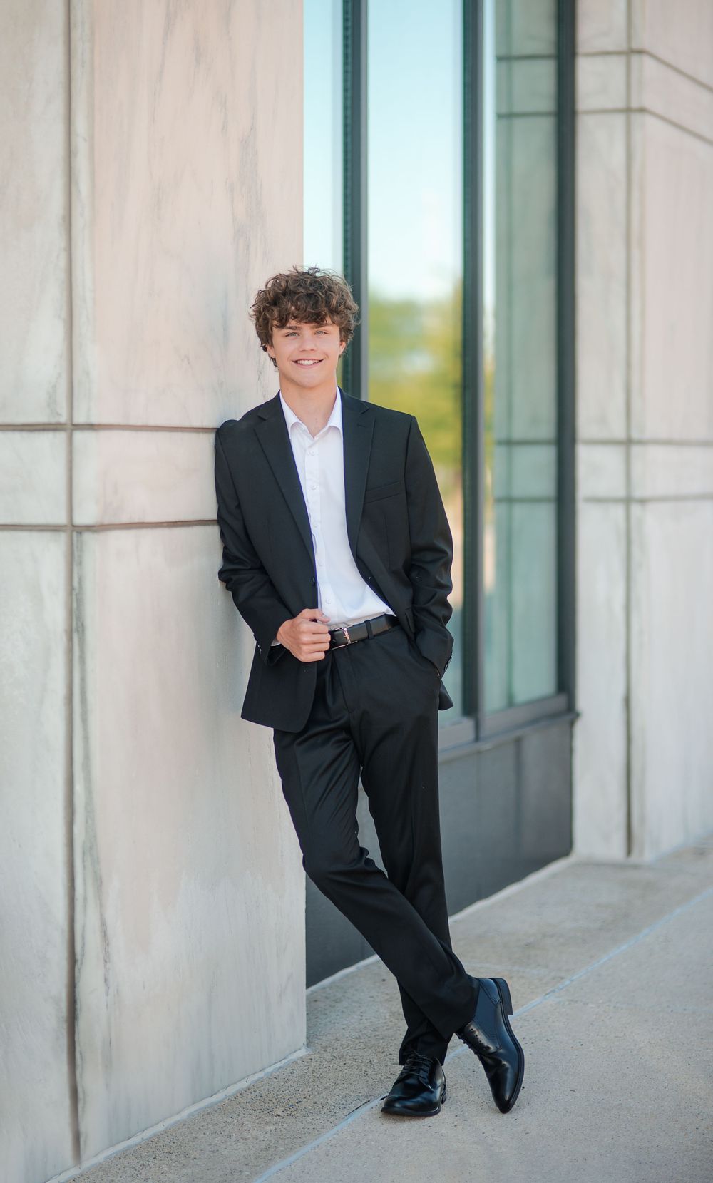 Young man in black suit and white shirt leaning against a stone wall, smiling confidently outdoors.