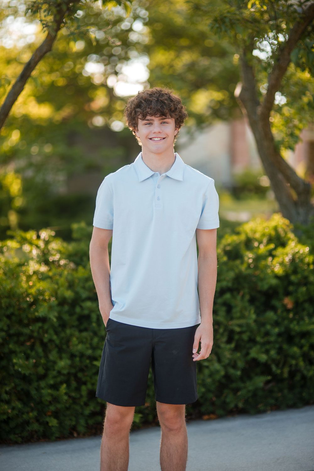 Smiling teenage boy with curly hair wearing a light blue polo shirt and dark shorts outdoors.