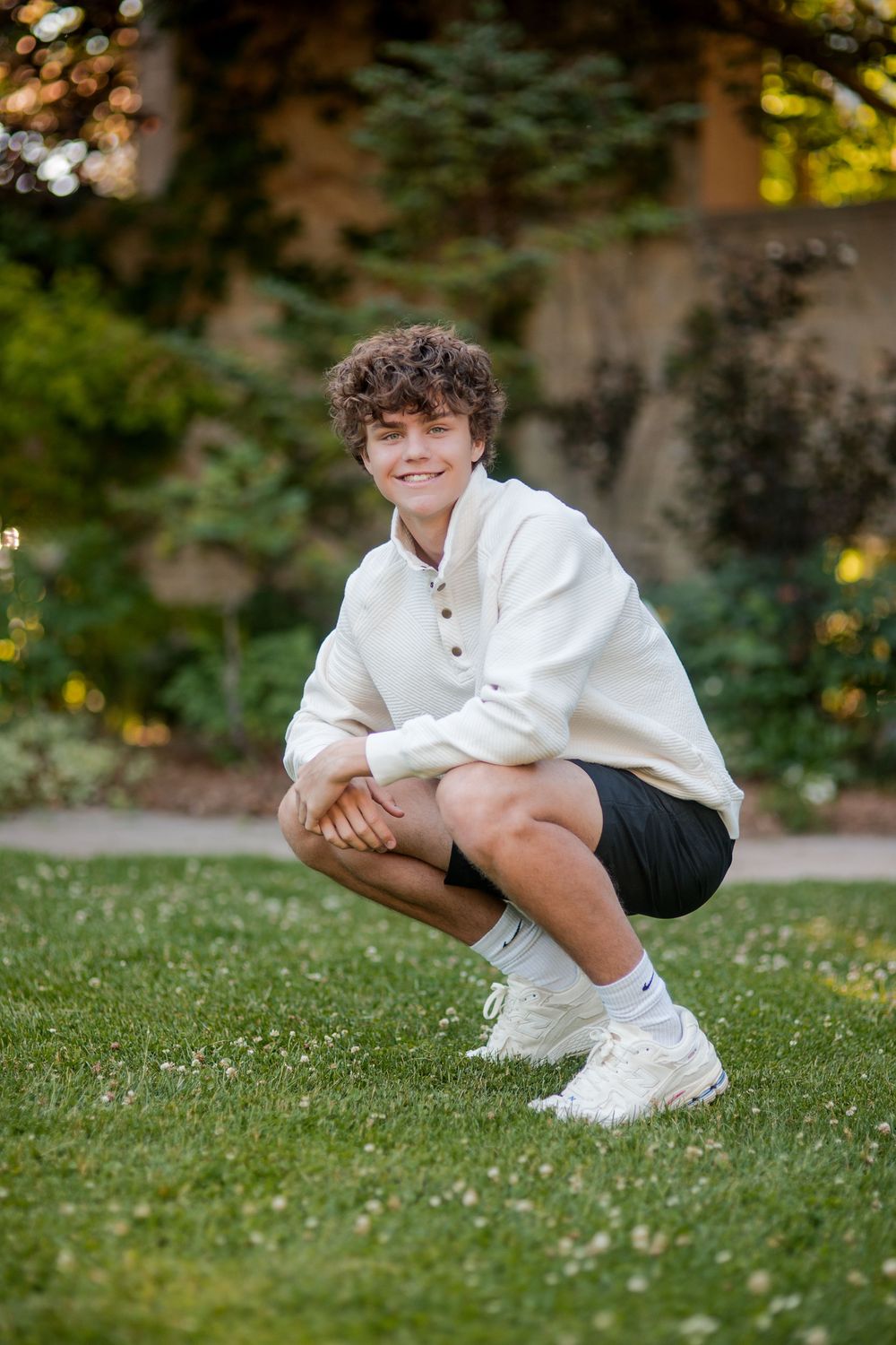 Young man in white sweater and black shorts crouching on green grass in a garden setting.