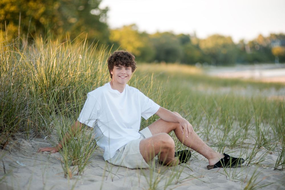 Smiling teenage boy sitting on sandy beach dunes wearing white shirt and shorts, surrounded by tall grass.