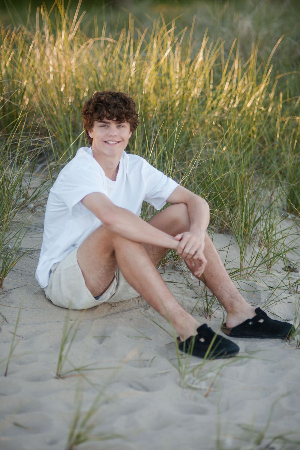 Teen boy with curly hair sits smiling on sandy beach dunes wearing white tee, khaki shorts, and black shoes.