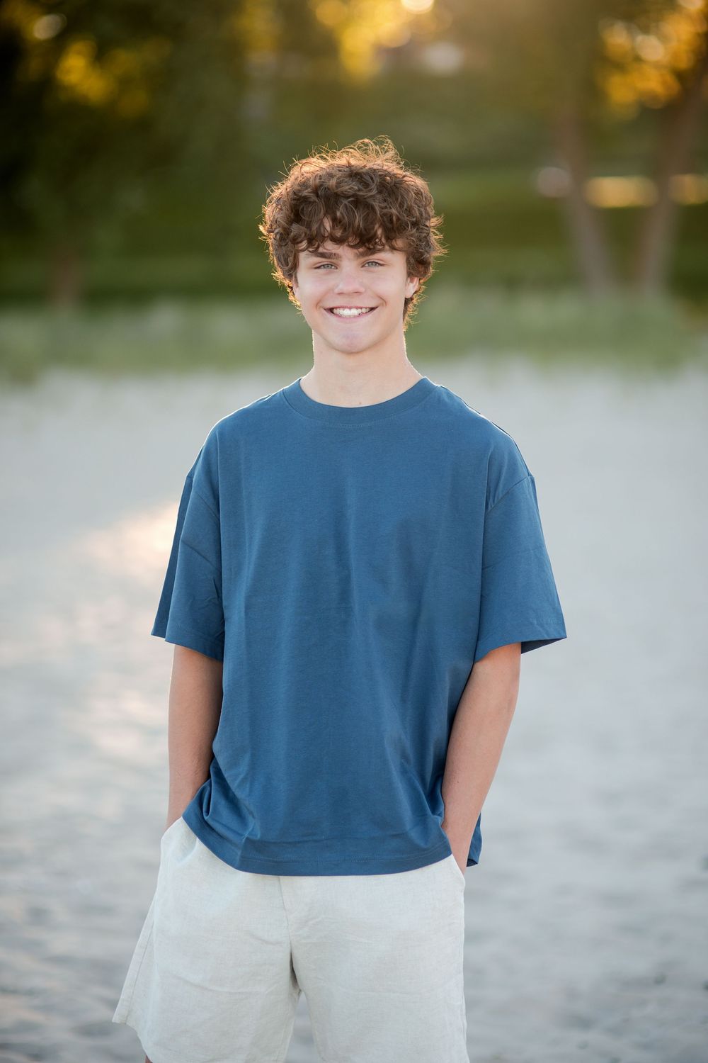 Smiling teenage boy with curly hair wearing a blue t-shirt and white shorts standing on a sandy beach.