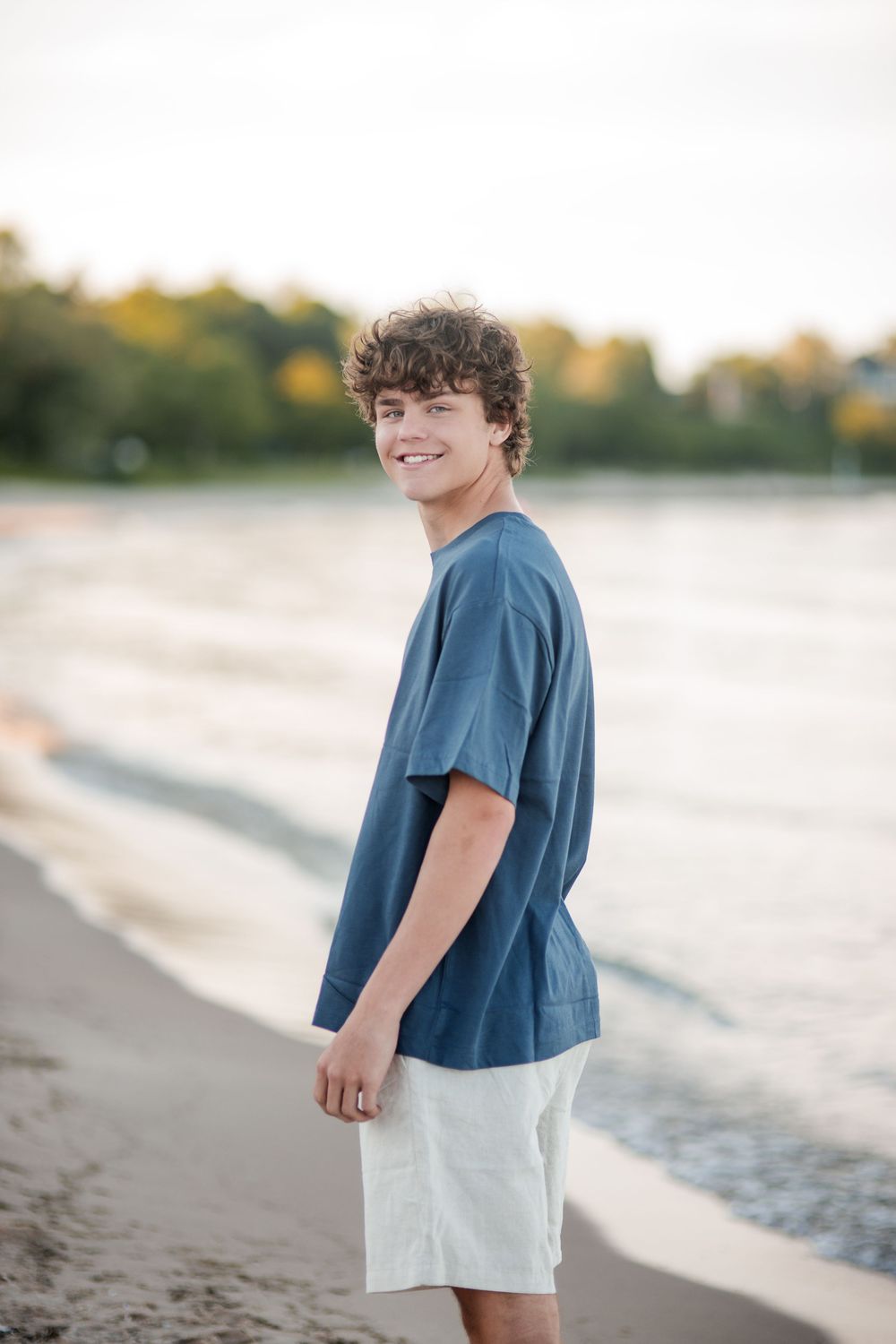 Smiling teenage boy in blue t-shirt and white shorts standing on a sandy beach near calm water.