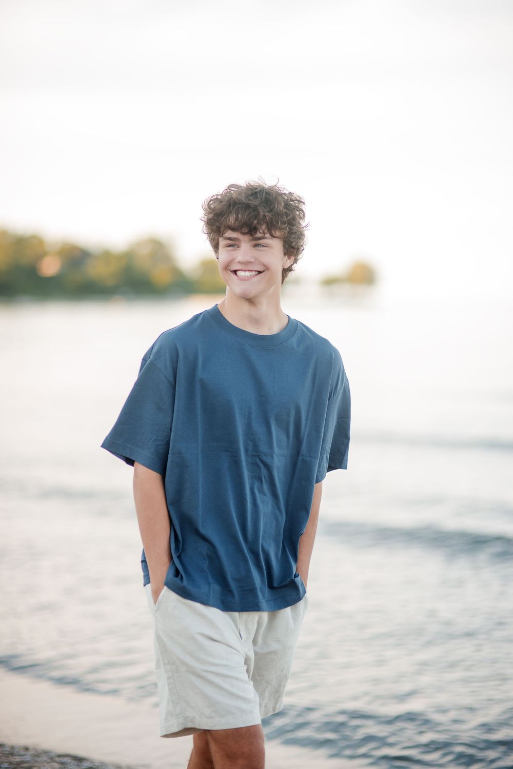 Smiling teenage boy with curly hair wearing a blue t-shirt and shorts standing on a beach near the water.