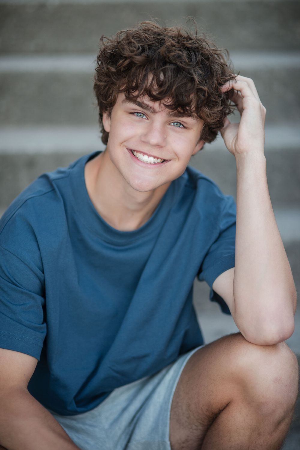 Smiling teenage boy with curly hair and blue eyes sits on steps, wearing a blue t-shirt and gray shorts.
