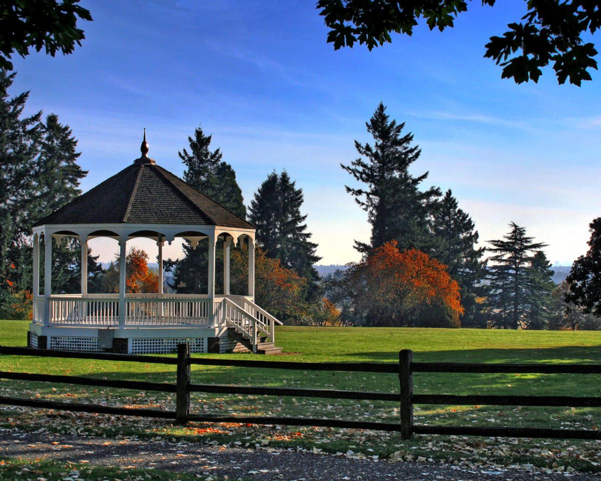 White gazebo in a lush green park with autumn trees and wooden fence under a clear blue sky.