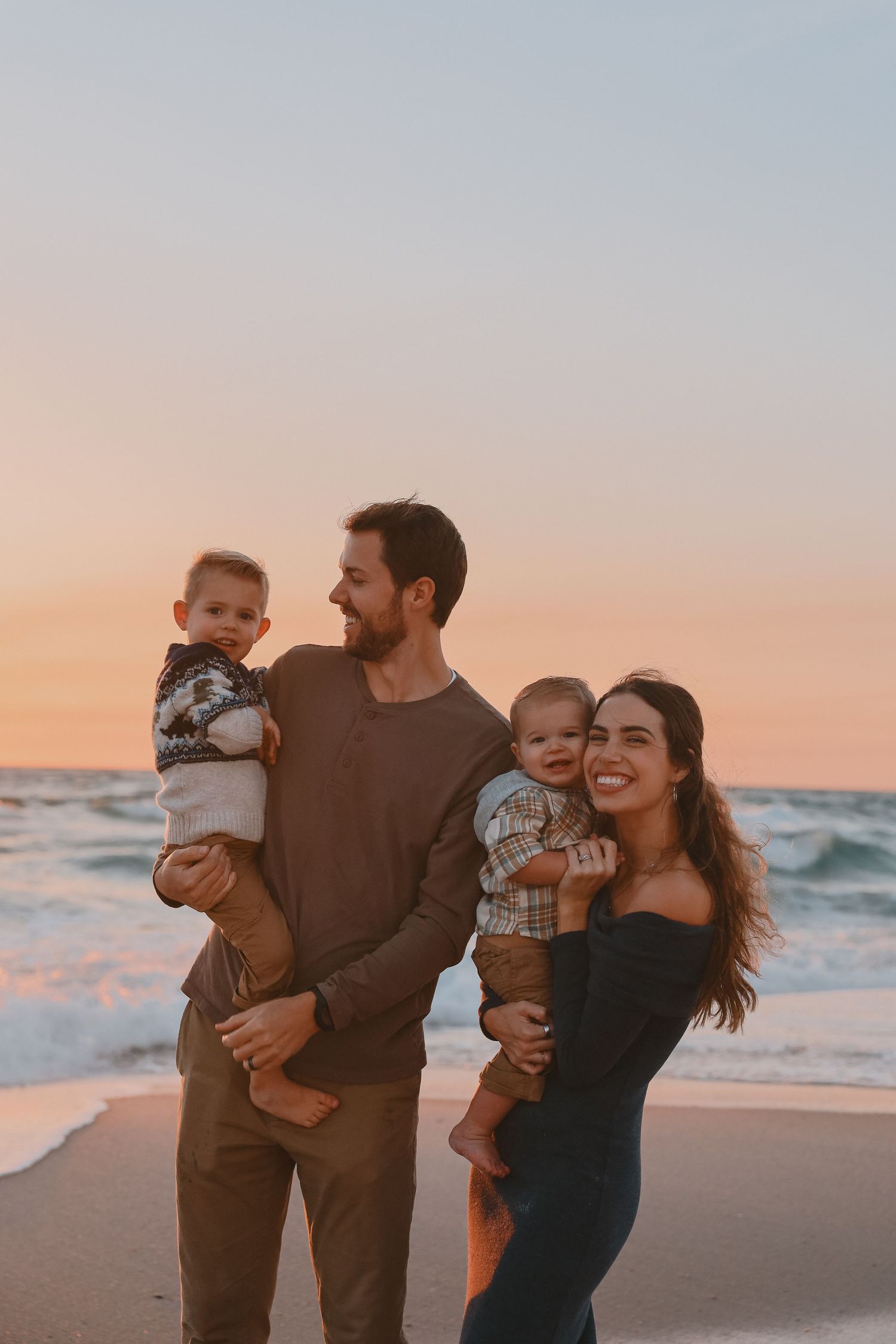 A happy family of four poses on a beach at sunset, with parents holding two toddlers as waves crash behind them.