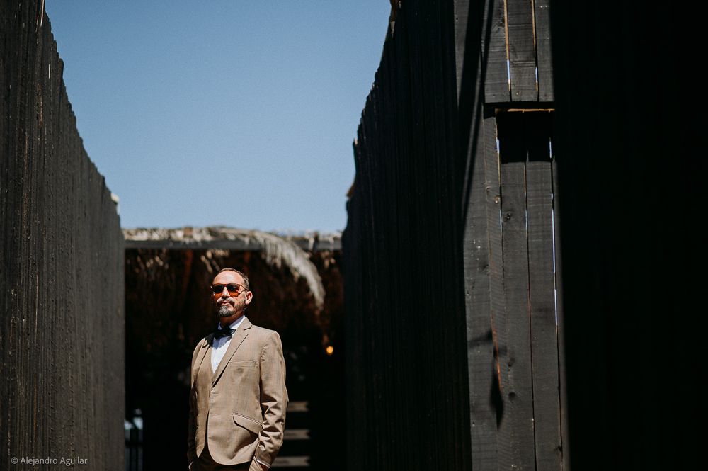 Man in beige suit and sunglasses stands between dark wooden walls in dramatic outdoor portrait.
