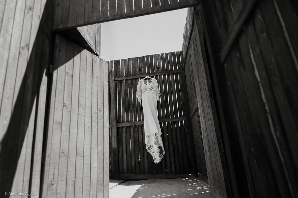 Black and white photo of a wedding dress hanging between rustic wooden walls under open sky.