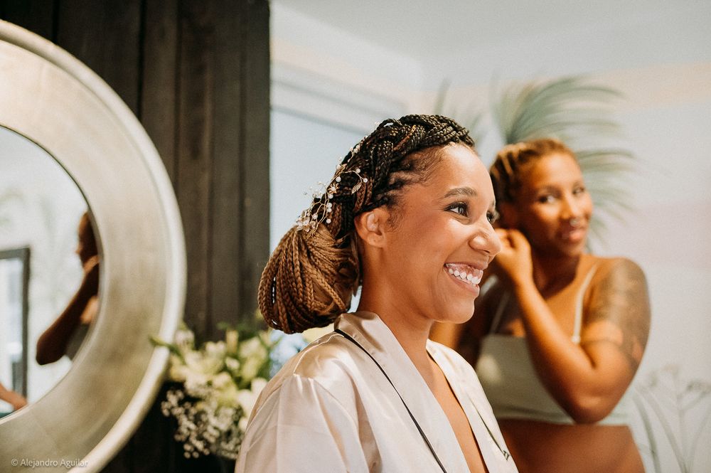 Bride smiling joyfully while getting hair styled with braids, bridesmaid adjusting earring in background.